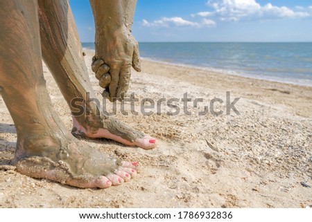Selective focus on beautiful female foot with bright red pedicure on the beach sand.  Hands smear healing mud on the ankles of the feet.  Skin rejuvenation while relaxing at sea. Spa. copy space. 