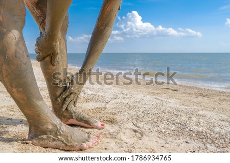 Selective focus on beautiful female foot with bright red pedicure on the beach sand.  Hands smear healing mud on the ankles of the feet.  Skin rejuvenation while relaxing at sea. Spa. copy space. 