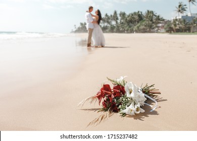 Selective focus on beautiful flowers bouquet in front of the wedding couple with the white beach background. Wedding bouquet in front of newlyweds wedding couple in background