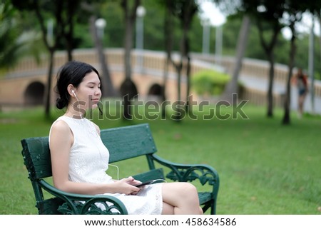 selective focus on a beautiful young lady wearing an earphone feel enjoy and relax while listening to music with her smartphone and blurry background of garden in the park
