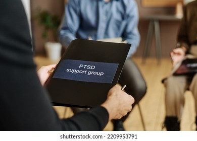 Selective Focus On Black Folder With Sticker Saying PTSD Support Group Held By Young Male Patient Of Psychotherapist Sitting In Front