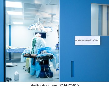 Selective Focus On The Blue Door With A Sign In Russian Language: Operating Room No. 1. Against A Blurred Background, Surgeons Operate On The Patient On The Operating Table.