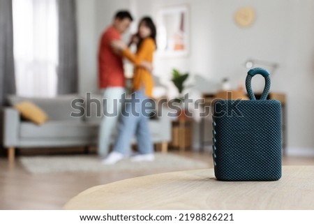 Selective focus on brand new portable speaker over dancing romantic asian couple, blurred background, copy space, home interior. Modern technologies and domestic entertainment concept