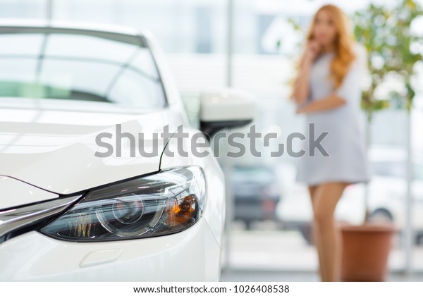 Selective focus on a car gorgeous elegant young\
woman choosing a new automobile at the dealership showroom on the\
background copyspace consumerism thinking decision buying cutomer\
driving.
