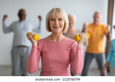 Selective Focus On Cheerful Senior Lady Posing With Fitness Tools, Multiracial Group Of Healthy Elderly People In Sportswear Doing Strength Building Workout With Dumbbells At Gym