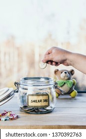 Selective focus on child hand, put euro coin in swear jar. Every time child curses or swears it has to put money as punishment in jar for safe keeps. Bad habit concept. Toy bear on the background.