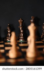 Selective focus on dark chess pieces lined up on a board against a black background, viewed from behind blurred light pieces in the foreground. Concept of tactical planning, strategy, and facing the opponent.