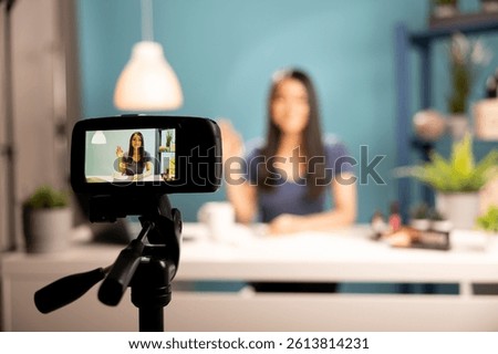 Selective focus on a device screen showing a smiling woman seated at desk, doing a hello hand gesture, vlogging in a studio. Digital camera on tripod records a beauty influencer during a livestream.