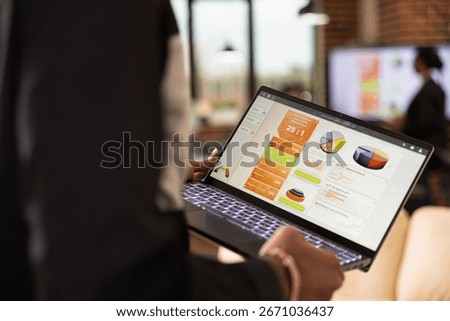 Selective focus on device screen displaying business analytics charts. Rearview closeup of african american person walking in office, reviewing financial graphs and evaluating kpi on laptop.