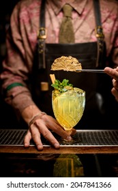 selective focus on dry yellow flower petal held with tweezers. Glass with bright yellow drink decorated with fresh mint leaves and bartender in the background