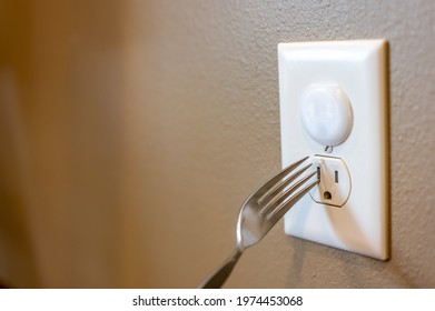 Selective Focus On Edge Of A Metal Fork About To Be Inserted Into An Open Electrical Outlet Socket. The Top Outlet Has A Plastic Insert For Childproofing Safety Protection.