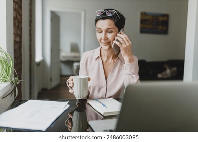 Selective focus on elegant successful wealthy entrepreneur having phone conversation with provider or supervisor during coffee break in office, sitting at table in front of laptop and documents - Powered by Shutterstock