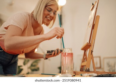 Selective focus on female creative artist's hand cleaning paintbrush in jar with water at art studio. Close up of talented female hipster painter cleaning brush in water while creating artwork. - Powered by Shutterstock