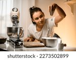 Selective focus on hand of woman pouring white flour into stainless bowl on table with modern kitchen appliances. Woman prepares dough pouring flour.