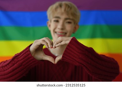 Selective focus on hands making heart shape by Young LGBT teenage boy with pride flag on the background, Support and celebrate the LGBT community.