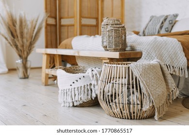 Selective Focus On Home Decor. Comfortable Bedroom In Bohemian Interior Style With Textile Sheet On Bed, Wooden Bench Seat, Bamboo Dressing Screen, Dry Plants In Vase, Wicker Basket