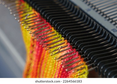 Selective focus on knitting machine needles. Openwork, colorful woolen scarf. Merino scarf on flat bed knitting machine. Simple knit fabric. Stockinette stitch - one of the most basic knitting pattens