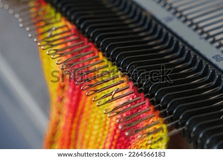 Selective focus on knitting machine needles. Openwork, colorful woolen scarf. Merino scarf on flat bed knitting machine. Simple knit fabric. Stockinette stitch - one of the most basic knitting pattens