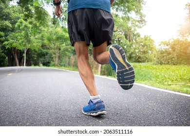 Selective Focus On The Left Foot And Leg. Man Legs Wearing Running Shoes Black Pants Are Jogging Exercises On The Road In The Park In The Morning. Jogging Strengthens The Body. The Background Is Green