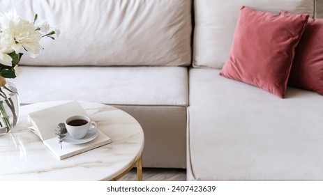 Selective focus on marble table with black coffee in with cup. Open book or diary journal with copy space paper pages close to vase with flowers next to comfortable couch with cushions in living room