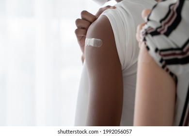 Selective focus on medicated plaster stuck on upper arm of dark skinned young man wear mask rolling up sleeves to show that he get vaccinated. Caucasian woman on blur foreground. White background - Powered by Shutterstock