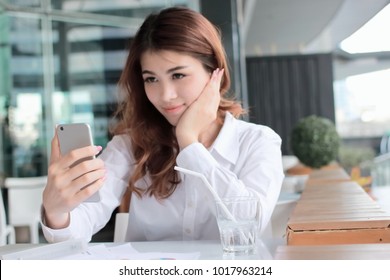 Selective Focus On Mobile Smart Phone In Hands Of Attractive Young Asian Woman Taking Photo Or Selfie In Office.