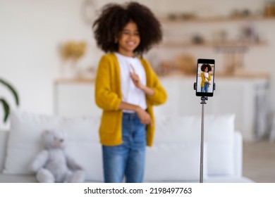 Selective Focus On Modern Cell Phone Set On Tripod Recording Video While Cute Curly Preteen  Girl In Casual Outfit Dancing, Having Fun Alone At Home, Blurred Background, Copy Space