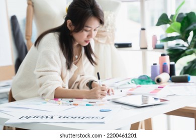 Selective Focus On Newspaper Placed On Desk Of Asian Beautiful Woman Fashion Designer Or Dressmaker Drawing Sketch Of New Collection Clothes On Paper, Working With Tablet On Desk At Workplace Studio