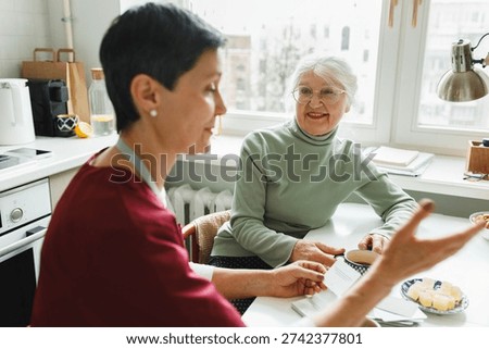 Selective focus on old smiling woman sitting at kitchen table near window and looking at her caregiver in red uniform, gesticulating explaining phrase, playing together charades to prevent dementia