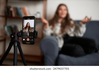 Selective focus on phone on tripod recording vlogger sitting on couch gesticulating and communicating with her followers, livestreaming for her blog in social media. Blogging as business - Powered by Shutterstock