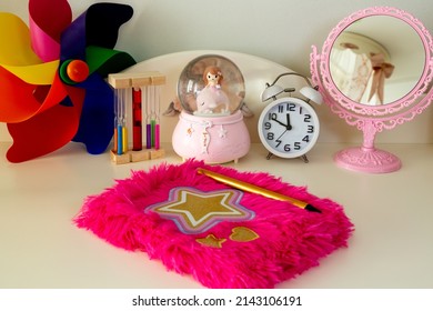 Selective Focus On Pink Ornate Diary On Top Of Girl's Bedroom Console. Background With Colorful Pinwheel, Decorative Hourglass, Classic Alarm Clock, Mermaid Glass Globe And Table Mirror.