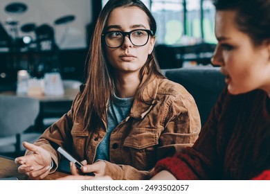 Selective focus on serious hipster girl in spectacles sitting college campus listening carefully female friend, two concentrated women studying together thinking on startup project using technology - Powered by Shutterstock