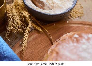 Selective Focus On The Spikelets Of Wheat Near A Blue Ceramic Bowl With White Flour And A Fresh Baked Whole Grain Wheaten Bread On Wooden Board, Cereal Grains Scattered On A Sackcloth. Overhead View