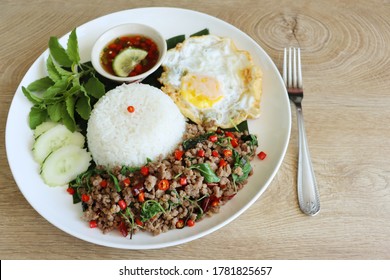 Selective focus on Thai food Stir fried minced pork with basil leaves(Pad Ka Prao) served with rice, fried egg, sliced cucumber,chilli in fish sauce and chopped basil on white plate with wooden table 