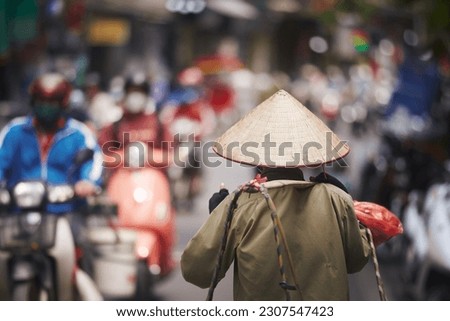 Selective focus on traditional conical hat of person walking against traffic motorbikes on busy street in Old Quarter in Hanoi, Vietnam.
