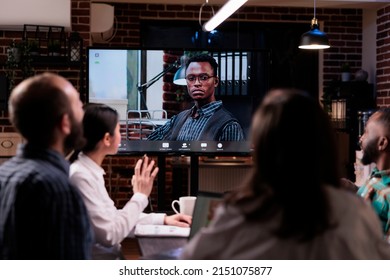 Selective Focus On Tv Screen With Serious Looking African American Business Owner In Online Video Conference At Office. Team In Late Night Meeting Working Overtime Greeting Ceo In Internet Call.