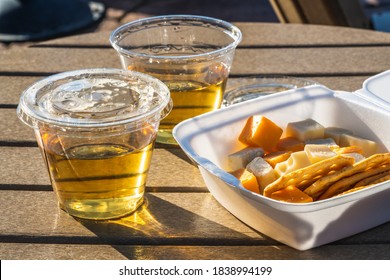 selective focus on a wine and cheese party in the time of Covid 19 with individualy packaged cheese and crackers and pre-portioned white wine in clear plastic cups outside in late afternoon sun