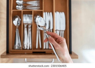 Selective focus on woman hand holding stainless steel spoon against blurred cutlery set in wooden organizer. Tidyness and neat in kitchen drawer. Keeping silverware clean concept