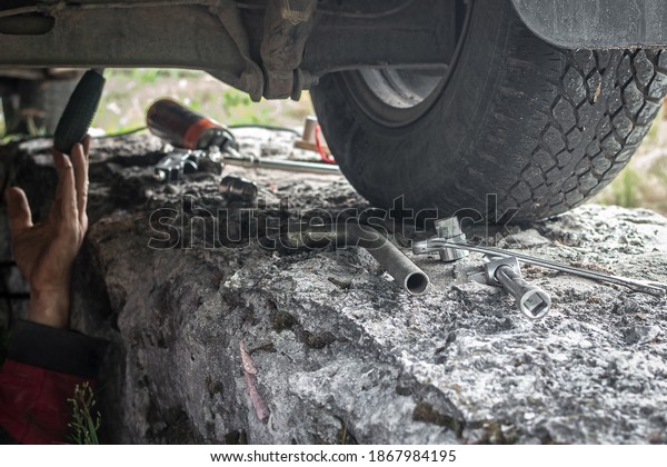 Selective focus on wrenches. A set of different\
wrenches lie on the concrete near the wheel of a car outdoors. The\
man lies under the car and makes repairs. Car bottom repair,\
exhaust system repair