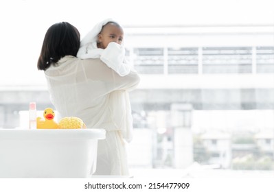 Selective Focus On Yellow Rubber Duck Toy, Sponge And Pink Shower Gel Bottle On White Baby Bathtub. Young Mother Wrap Up Cold African Newborn Baby Warm In Towel In Embrace After Bathing At Bathroom