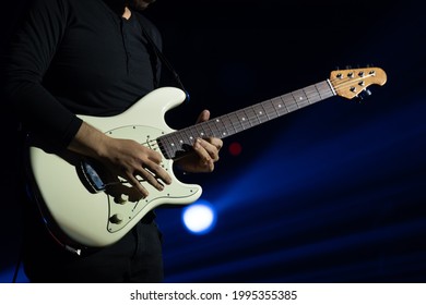 Selective focus on young Asian man guitarist's hand performing playing lead electric guitar solo with full spectrum of light and sound at concert. Blue light background.