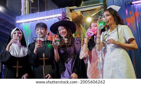 Selective focus on young Asian woman dressed as witch smiling and holding drink at Halloween party with group of friends having fun dressed up as priest, nun, victim and nurse at club