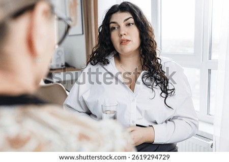 Selective focus on young plus size woman with curly hair and glass of water in her hands consulting with her therapist, trying to solve her mental issues. Psychotherapy support concept