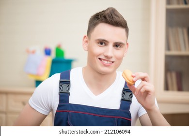 Selective focus on young smiling dark-haired janitor wearing white T-shirt and blue overalls eating tasty cookie during his break. Blue pail with cleaners standing on the commode on background