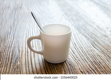 Selective focus of one white empty coffee cup with silver spoon on a rustic oak wooden table with bright background.