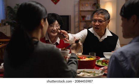 selective focus with over shoulder view smiling asian elderly couple grandfather and grandmother holding food with chopsticks and giving to their daughter in law at dinner table on eve of new year