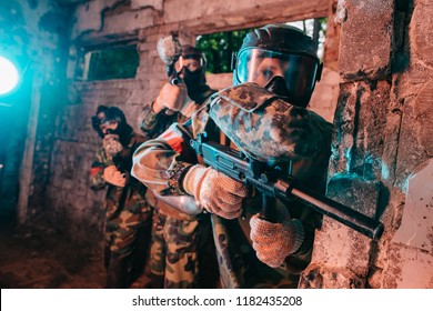 selective focus of paintball team in uniform and protective masks playing paintball with marker guns in abandoned building