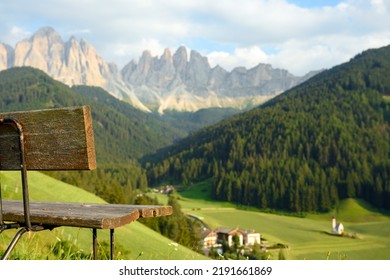 (Selective focus) Park bench in the foreground with defocused Church of St. John (San Giovanni in Ranui) that stands out in the green meadows, in the heart of the beautiful Dolomitic landscape.
