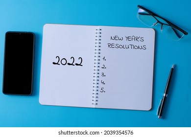 Selective focus of pen, glasses, smartphone and notebook written with 2022 new year's resolutions on blue background.