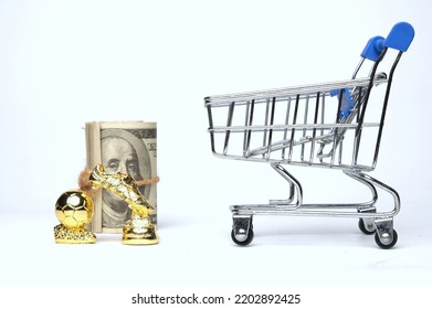 Selective Focus Picture Of Shopping Cart, Golden Ball And Boot Trophy Miniature With Fake Money Insight. Football Player Transfer Window Concept.